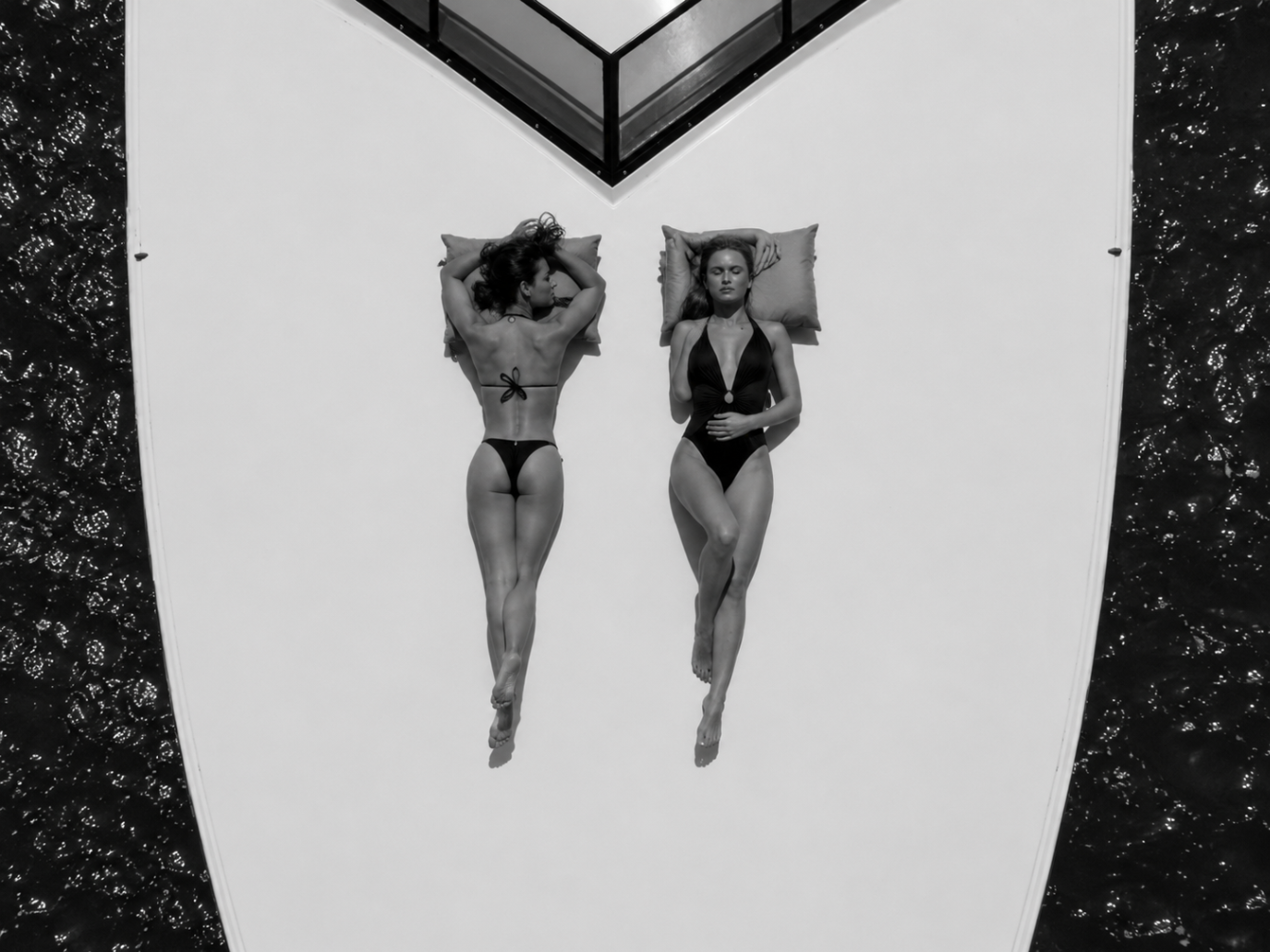 Aerial view of two women resting on white cushions in a symmetric layout above dark water