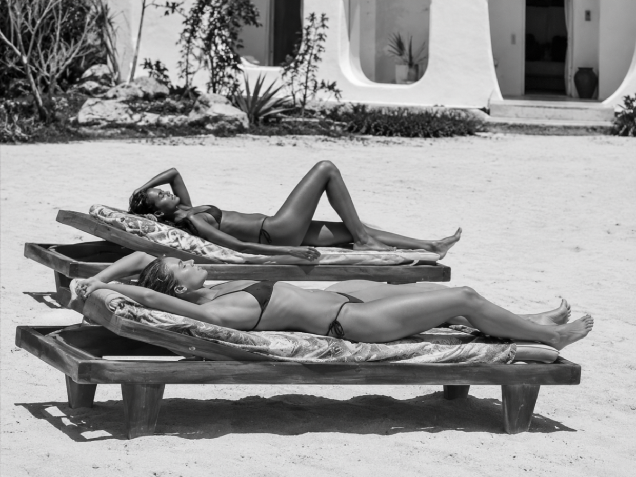 Two women sunbathing on wooden loungers outside a white villa