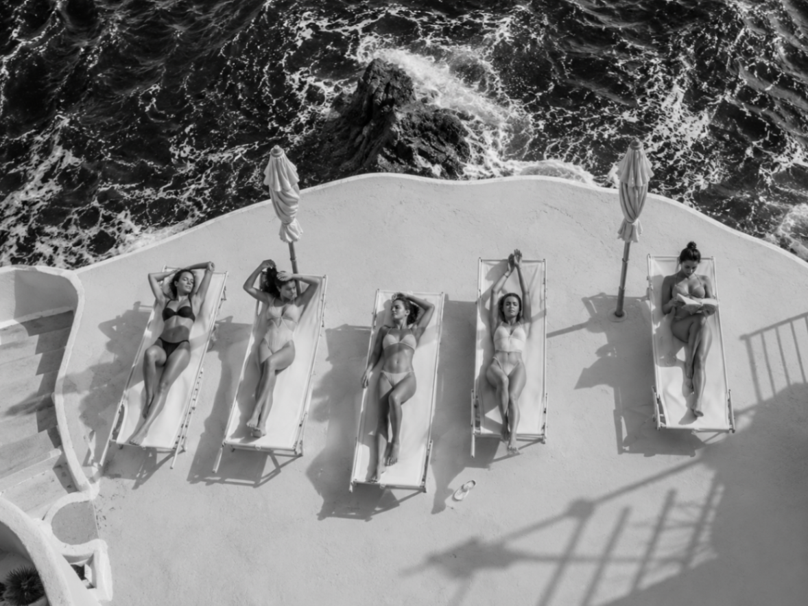 Aerial view of five women on loungers above the sea, evoking decades of ease