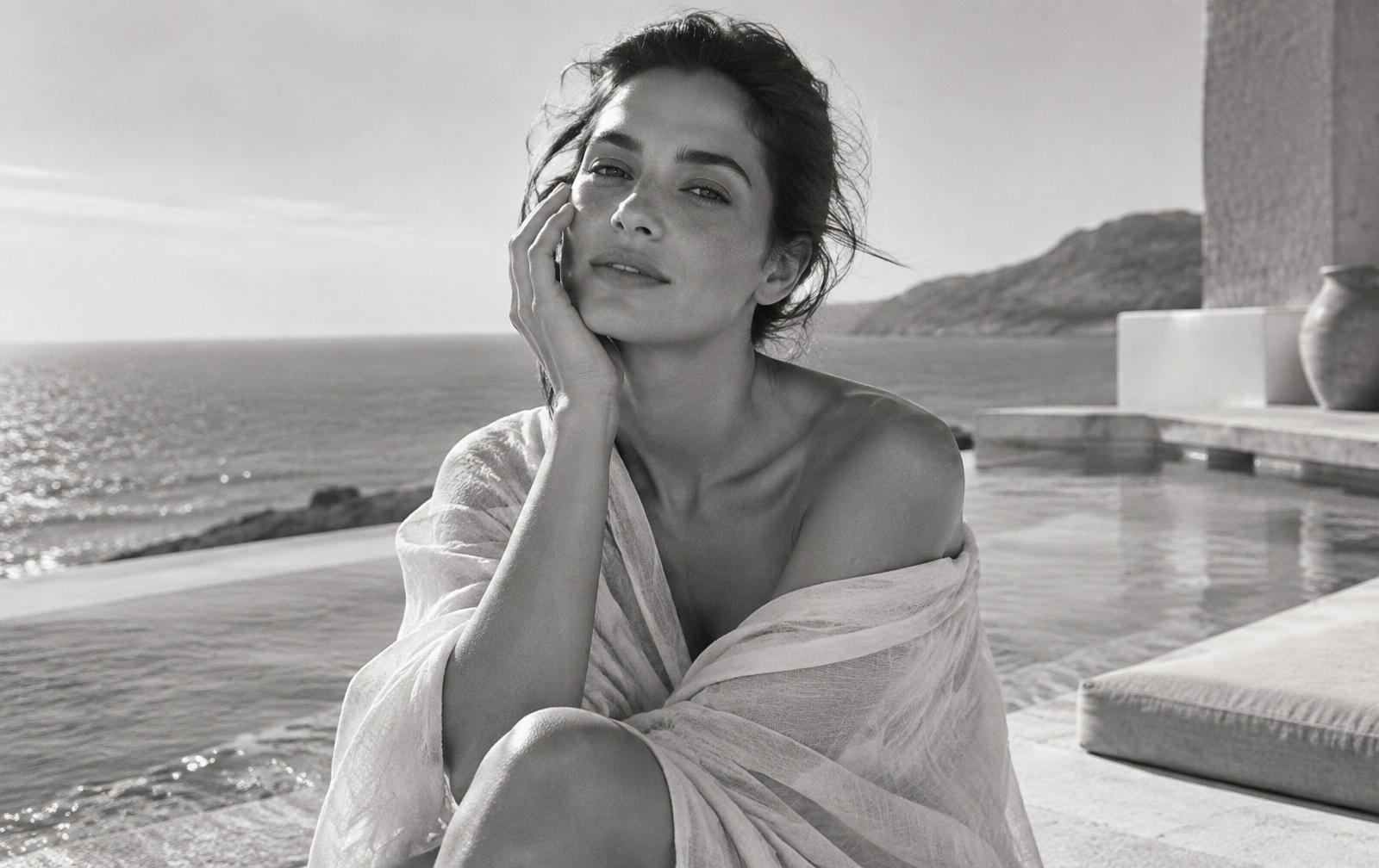 Black-and-white editorial portrait of a woman beside a coastal pool, gently touching her cheek