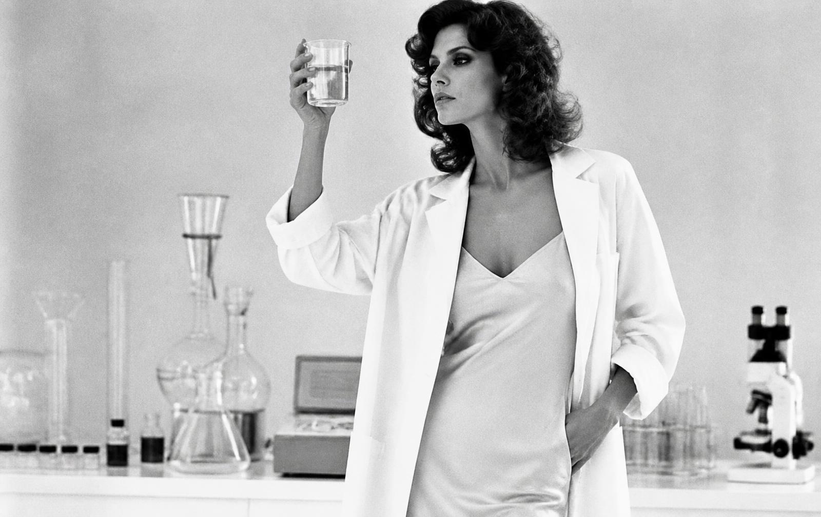Black-and-white editorial photo of a woman in a lab coat holding a beaker of serum to the light