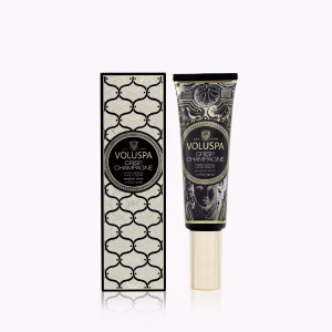 A Voluspa Crisp Champagne Hand Cream tube stands beside its decorative black and gold box, both displaying elegant labels, set against a white background.