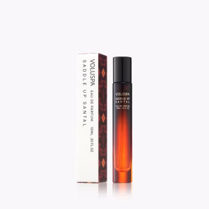 A bottle of Voluspa Saddle Up Santal Perfume is displayed beside its matching rectangular box, both in sleek brown and white packaging with minimalist branding.