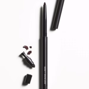 A Revitalash Defining Liner–slate, with its black retractable pencil and cap off, lies on a white surface. Small liner pieces and a smudger are beside it. "DEFINING LINER" is printed on the pencil.