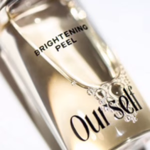 A close-up of a clear bottle labeled "Ourself Brightening Peel," featuring a light-colored liquid with visible bubbles inside, sits on a white surface.