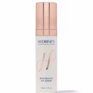 A white bottle with a rose gold cap labeled Hydrinity Restorative HA Serum, featuring a large stylized H on the front.