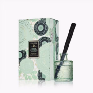 The Voluspa White Cypress Reed Diffuser, featuring black reeds in a decorative glass vessel, is displayed next to its matching box adorned with green and black floral patterns and a "White Cypress" label.