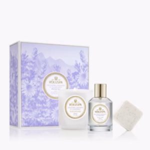 The Voluspa Mountain Lavender & Chamomile Gift Set includes a lavender floral box, a white candle, a clear bottle with a gold cap, and a square soap, set against a light background.