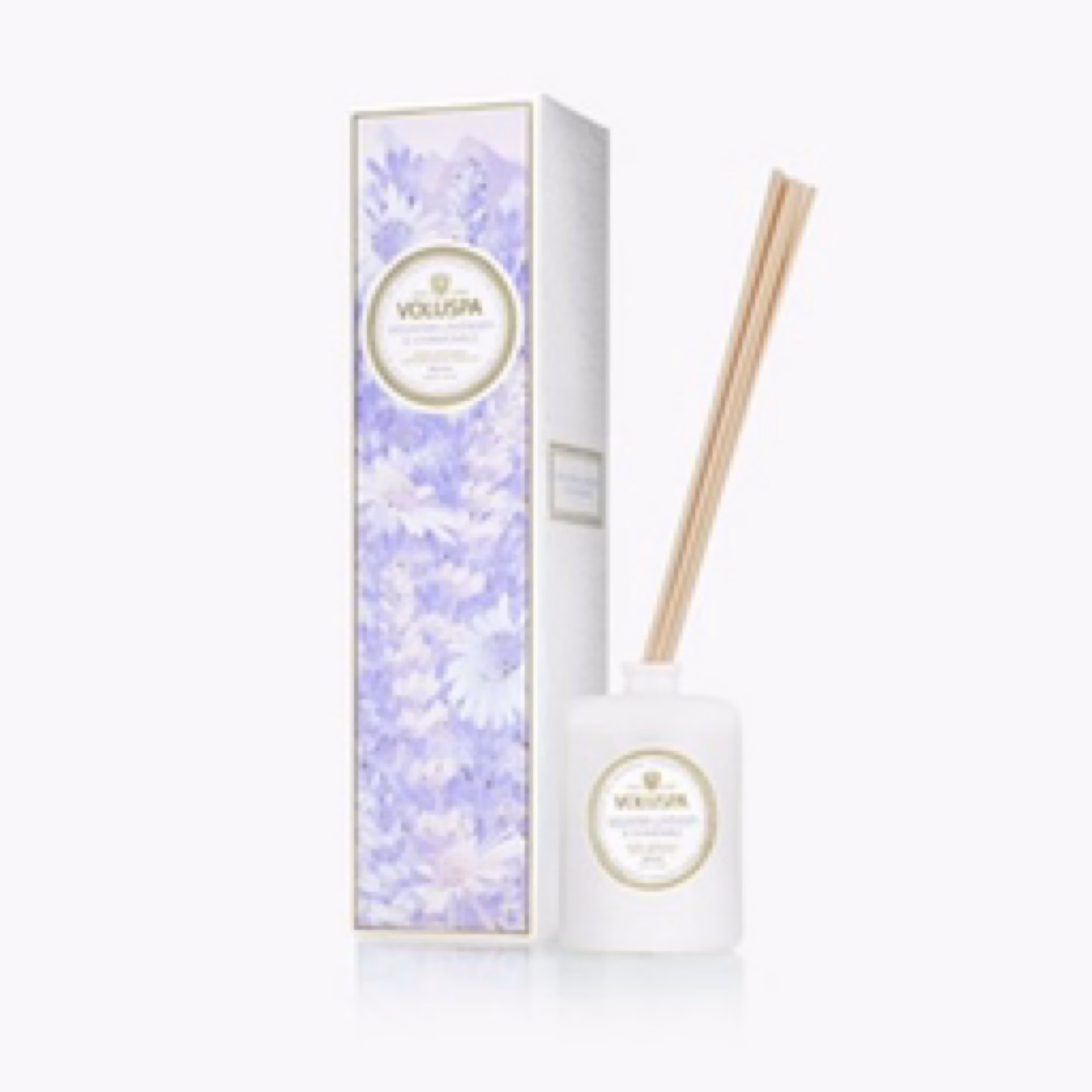 The Voluspa Mountain Lavender & Chamomile Reed Diffuser features a white bottle with natural reeds, shown beside its decorative box adorned with purple floral designs on a white background.