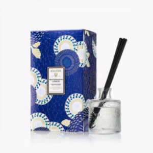 A clear glass diffuser bottle with black reeds is displayed beside a decorative blue box featuring white, gold, and teal circular patterns. The box label reads Voluspa Mediterranean Lemon Reed Diffuser.