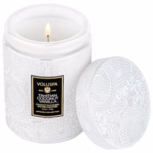 A lit Voluspa Tahitian Coconut Vanilla Candle Small 5.5oz jar sits in a white embossed container, featuring gold and black label details and an ornate matching lid placed beside it.